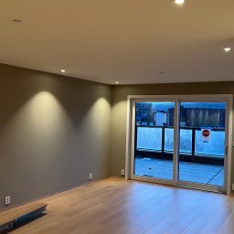 SPONTANDEKOR Tomasz Antoniewicz - Wnętrze po malowaniu: ściany w odcieniach beżu, jasna podłoga, przesuwne drzwi balkonowe z widokiem na zewnątrz. Na podłodze leżą panele. Oświetlenie punktowe.