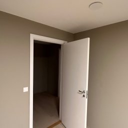 SPONTANDEKOR Tomasz Antoniewicz - Pomalowane na beżowo wnętrze z otwartymi, białymi drzwiami. Widoczna podłoga z jasnego drewna i biała oprawa oświetleniowa na suficie. Minimalistyczny design.