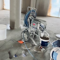 SPONTANDEKOR Tomasz Antoniewicz - Agregat malarski po użyciu, z widocznymi śladami farby, stojący na podłodze w trakcie remontu. Obok puszki z farbą i narzędzia malarskie.