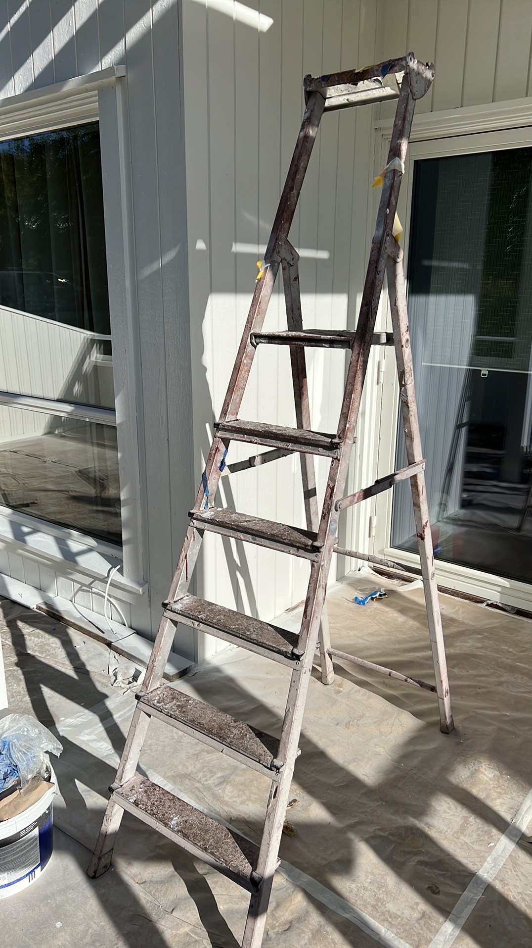 Drabina malarska oparta o białą elewację budynku z widocznymi oknami. Podłoże zabezpieczone folią malarską. Ślady farby na drabinie.