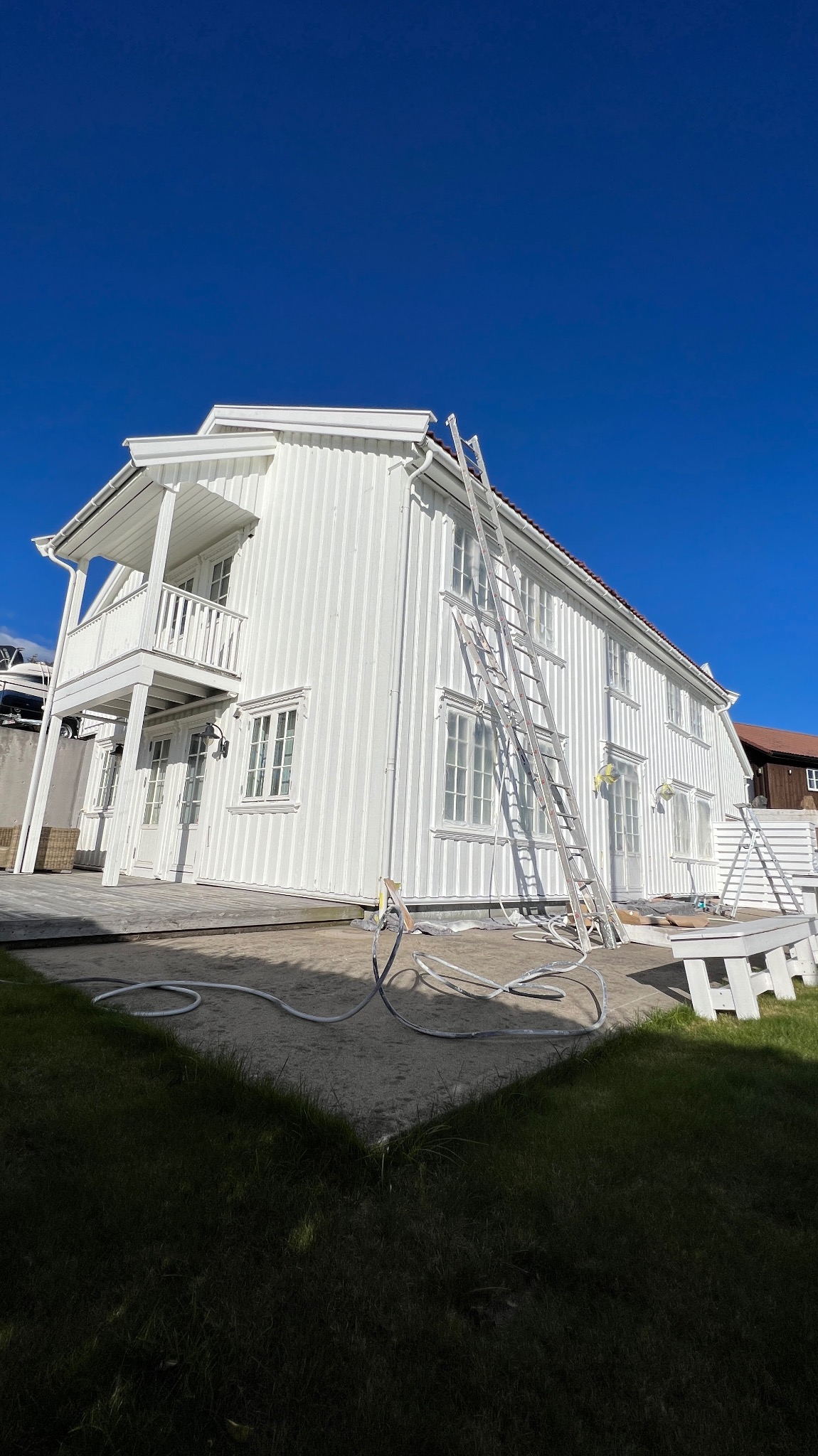 Biały, dwukondygnacyjny dom z drewnianą elewacją po malowaniu, z drabiną opartą o ścianę. Widoczne zabezpieczenia okien i węże malarskie. Błękitne niebo w tle.