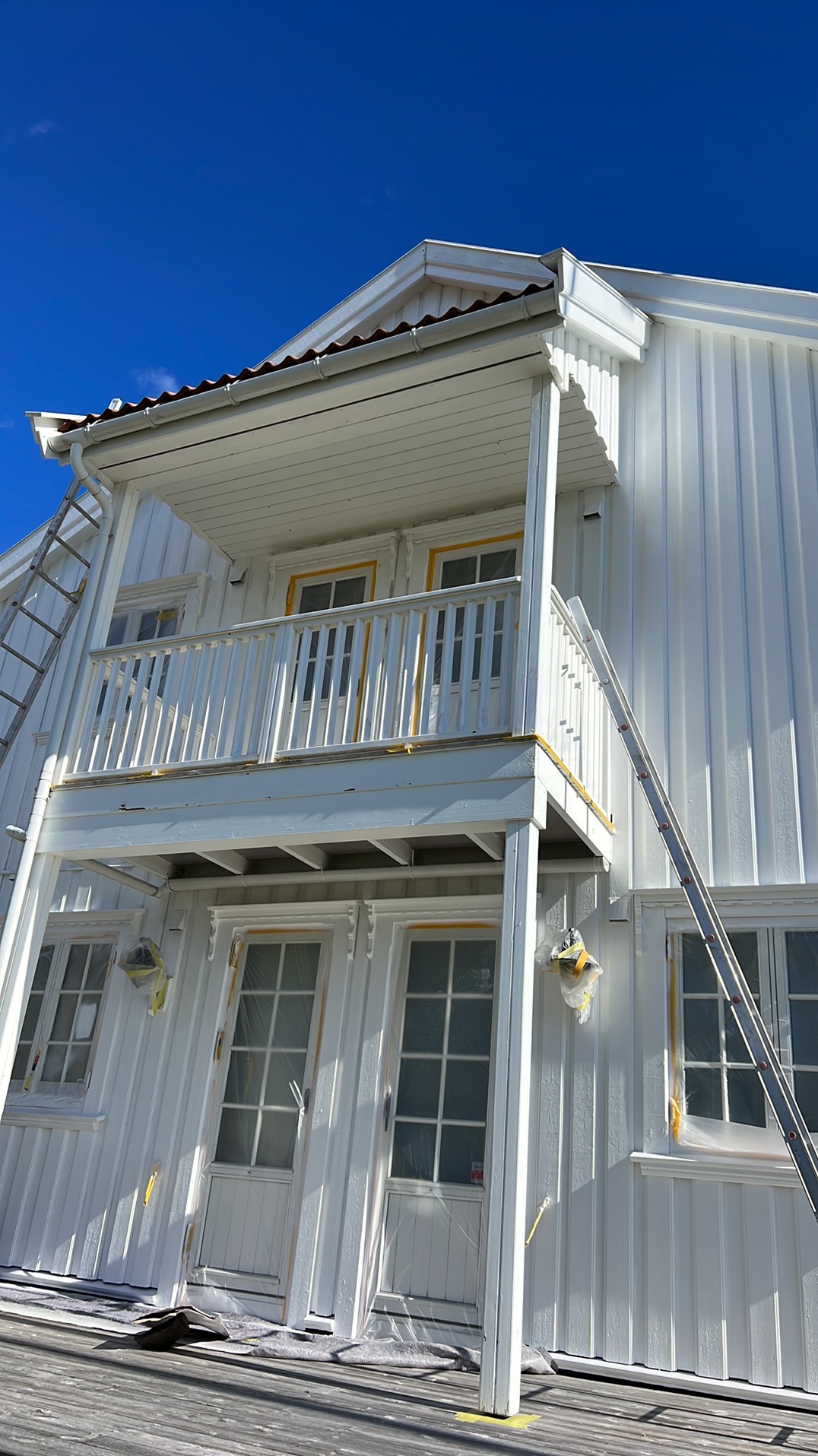 Biała elewacja domu z balkonem w trakcie malowania, zabezpieczone okna folią, drabina oparta o ścianę, błękitne niebo w tle. Widok z dołu.