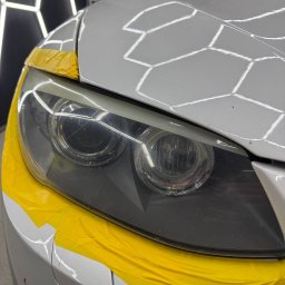 King Art - Biały samochód z reflektorami zabezpieczonymi żółtą taśmą przed polerowaniem. Widoczne zarysowania na szkle reflektorów. Oświetlenie warsztatowe w tle.