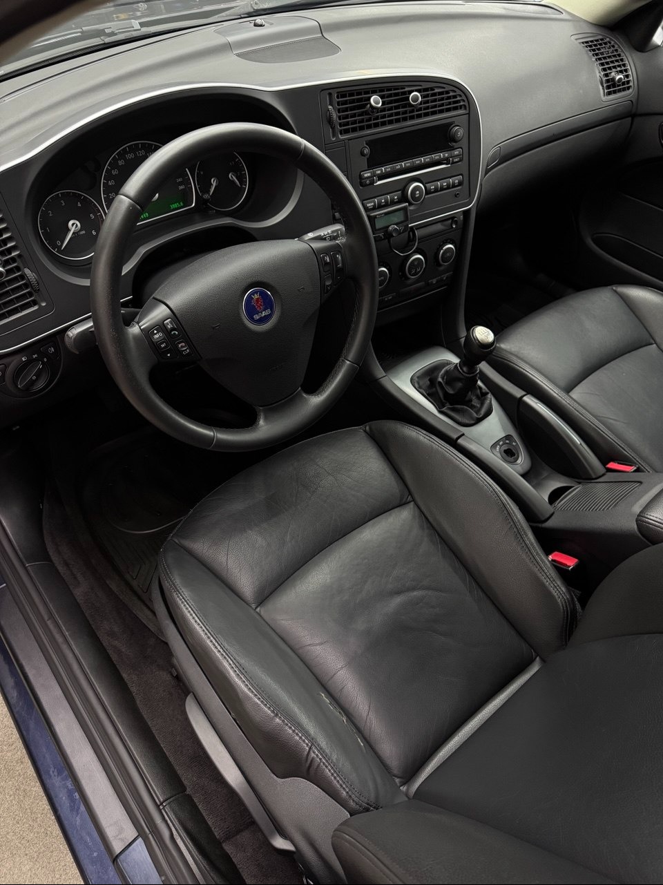 Wnętrze samochodu Saab 9-3 z czarną skórzaną tapicerką. Widoczna kierownica z logo, deska rozdzielcza i przednie fotele. Ciemne wykończenie wnętrza.