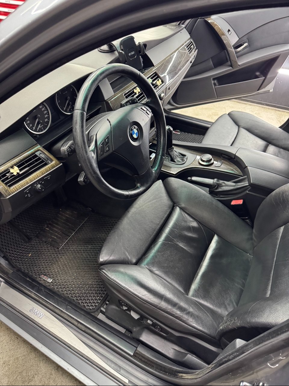 Wnętrze samochodu BMW z czarną skórzaną tapicerką, widoczne fotele, kierownica i deska rozdzielcza. Dywaniki gumowe. Widoczne ślady użytkowania na fotelu kierowcy.