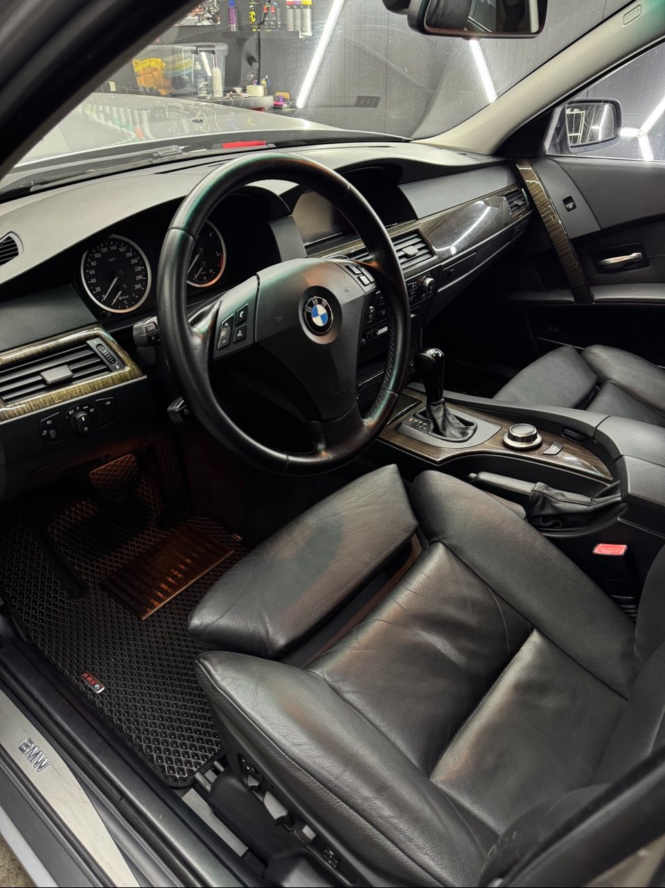 Wnętrze BMW po detailingu: czarne skórzane fotele, deska rozdzielcza z drewnianymi akcentami, widoczne dywaniki z logo BMW. Oświetlenie studyjne podkreśla detale.