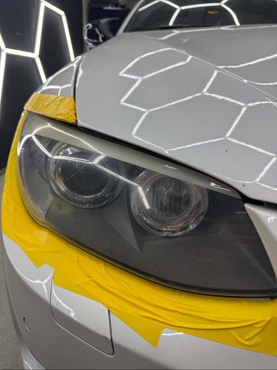 Biały samochód z reflektorami zabezpieczonymi żółtą taśmą przed polerowaniem. Widoczne zarysowania na szkle reflektorów. Oświetlenie warsztatowe w tle.