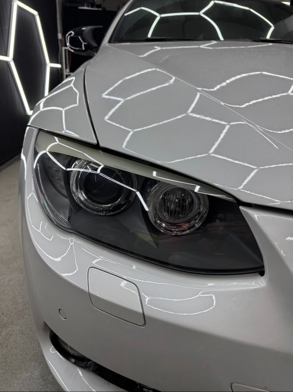 Biały samochód BMW po detailingu, reflektory i maska z odbiciami lamp w kształcie plastra miodu. Widok z bliska na przód pojazdu, czysty i błyszczący lakier.