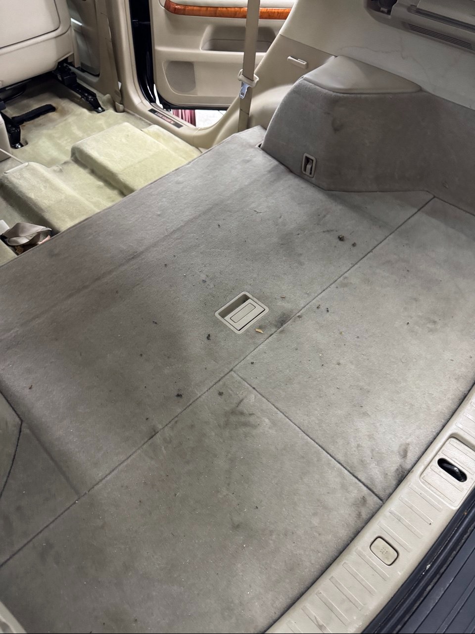 Brudna, beżowa tapicerka bagażnika samochodu przed praniem. Widoczne plamy i zabrudzenia na powierzchni materiału. Ujęcie z góry, wnętrze pojazdu.