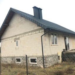 DEKARMAX Jakub Olbryś - Niedokończony dom z ciemną dachówką i rynnami, widoczne okna i komin. Budynek w stanie surowym, otoczony siatką i suchą trawą. W tle las i pochmurne niebo.