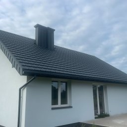 DEKARMAX Jakub Olbryś - Dom jednorodzinny z ciemną dachówką i kominem w kolorze dachu. Widoczne okna i drzwi balkonowe. Tło stanowi błękitne niebo z chmurami i zieleń drzew.