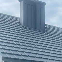 DEKARMAX Jakub Olbryś - Grafitowy dach z blachodachówki z kominem wentylacyjnym w nowoczesnym stylu. Widoczne orynnowanie. Ujęcie z dołu, pochmurne niebo w tle.