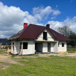 DEKARMAX Jakub Olbryś - Dom w trakcie budowy z częściowo ukończonym, brązowym dachem z blachy trapezowej i czerwonym kominem. Widoczne rusztowania i elementy konstrukcyjne.