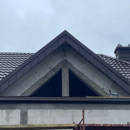 DEKARMAX Jakub Olbryś - Fragment dachu z brązową blachodachówką i kominem w Jednorożcu, widok z dołu na szczyt ściany z pustaków. Widoczne rusztowanie i elementy konstrukcyjne.