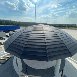 DEKARMAX Jakub Olbryś - Nowoczesna altana z ciemnoszarą dachówką, widok z góry na tle błękitnego nieba i zieleni. Nietypowy kształt dachu i jasne kolumny altany.