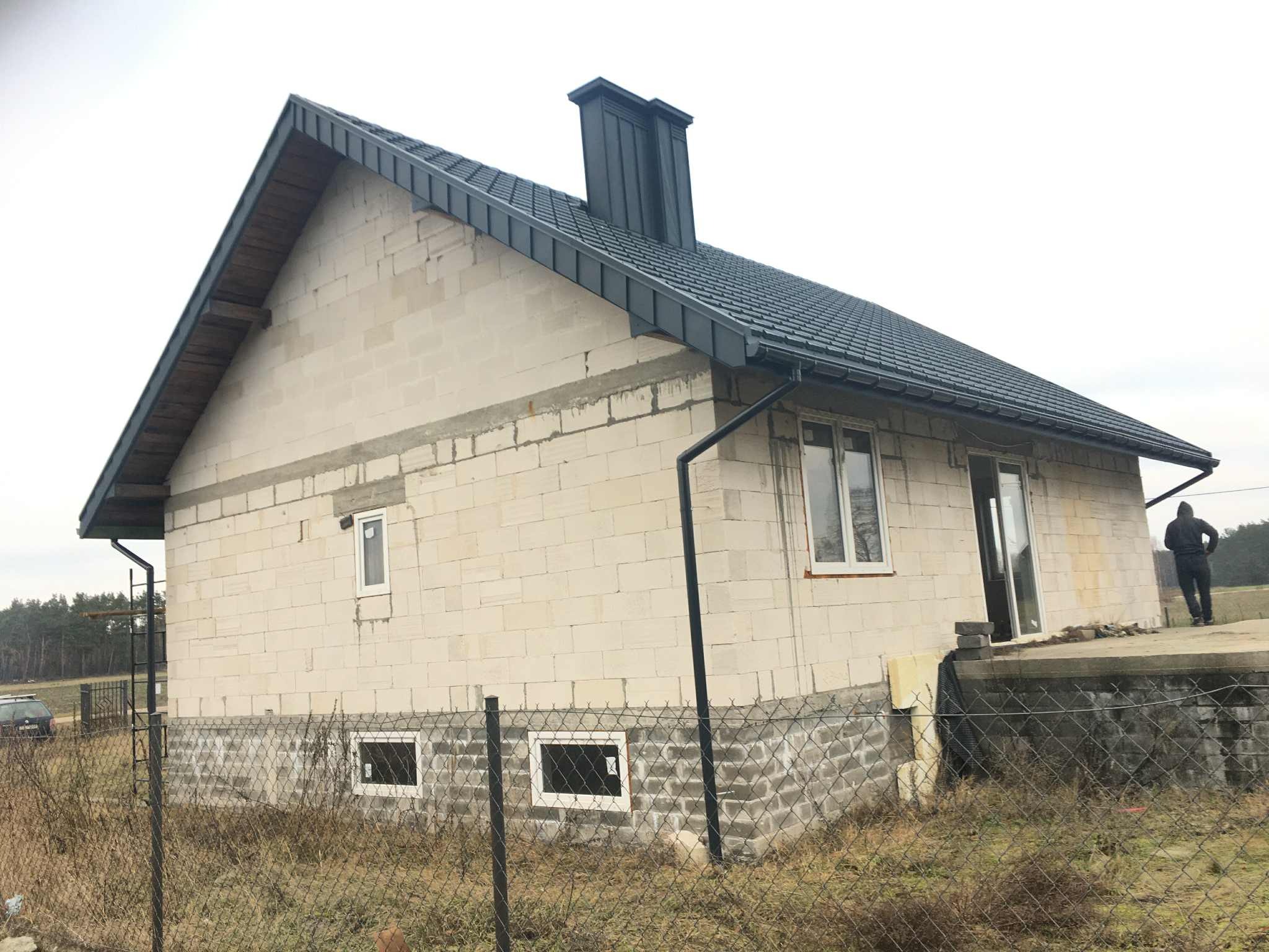 Niedokończony dom z ciemną dachówką i rynnami, widoczne okna i komin. Budynek w stanie surowym, otoczony siatką i suchą trawą. W tle las i pochmurne niebo.