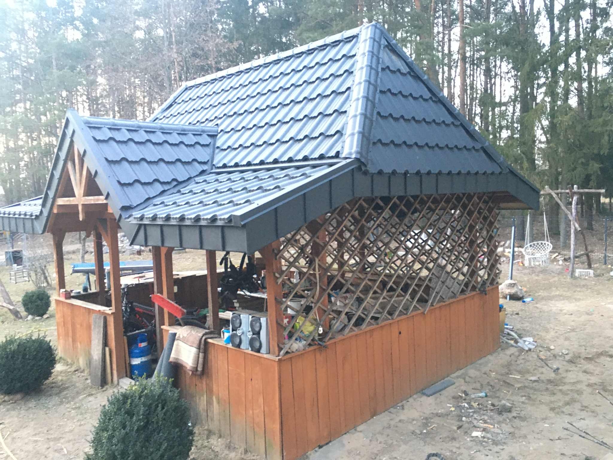 Zadaszona altana ogrodowa z nowym, ciemnoszarym dachem z blachodachówki, widoczna z boku na tle drzew. Konstrukcja drewniana, częściowo osłonięta kratownicą.