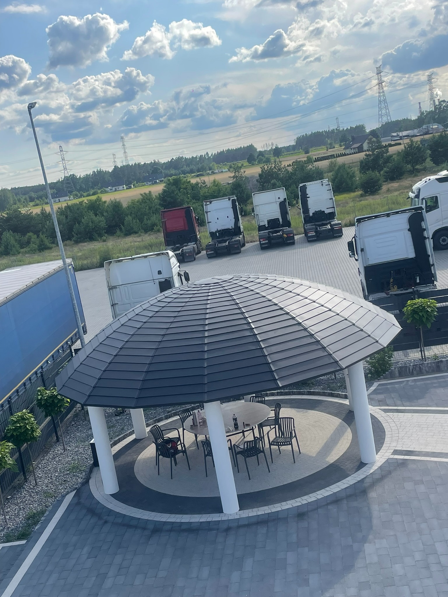 Altana z dachem z blachodachówki w nowoczesnym stylu, widok z góry. W tle zaparkowane samochody ciężarowe i słupy wysokiego napięcia.