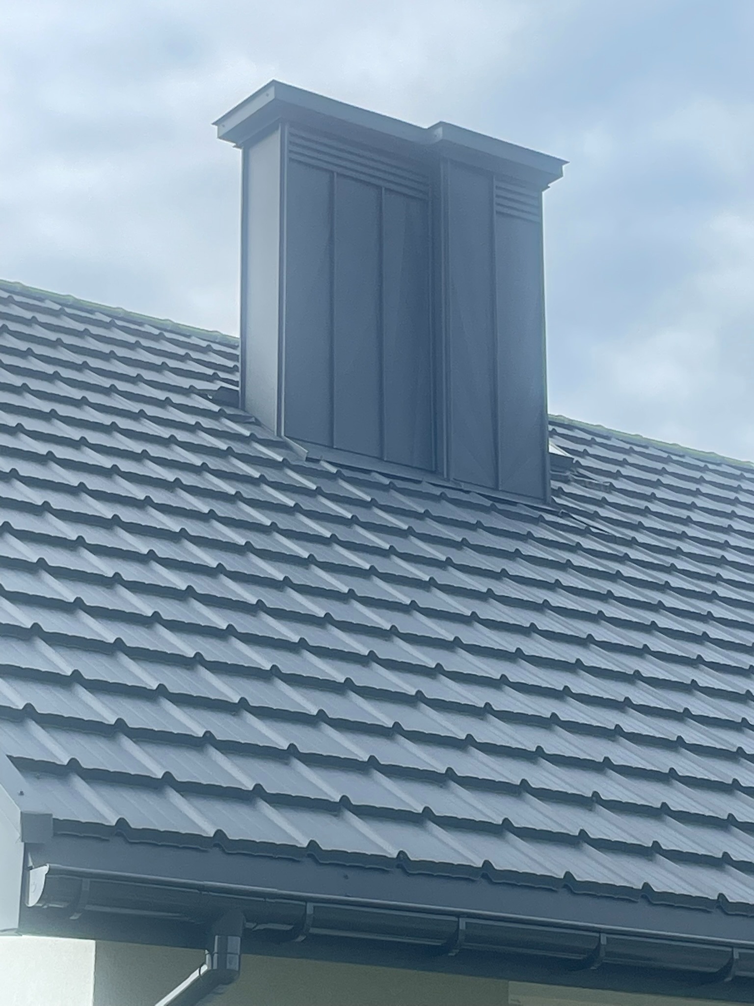 Grafitowy dach z blachodachówki z kominem wentylacyjnym w nowoczesnym stylu. Widoczne orynnowanie. Ujęcie z dołu, pochmurne niebo w tle.