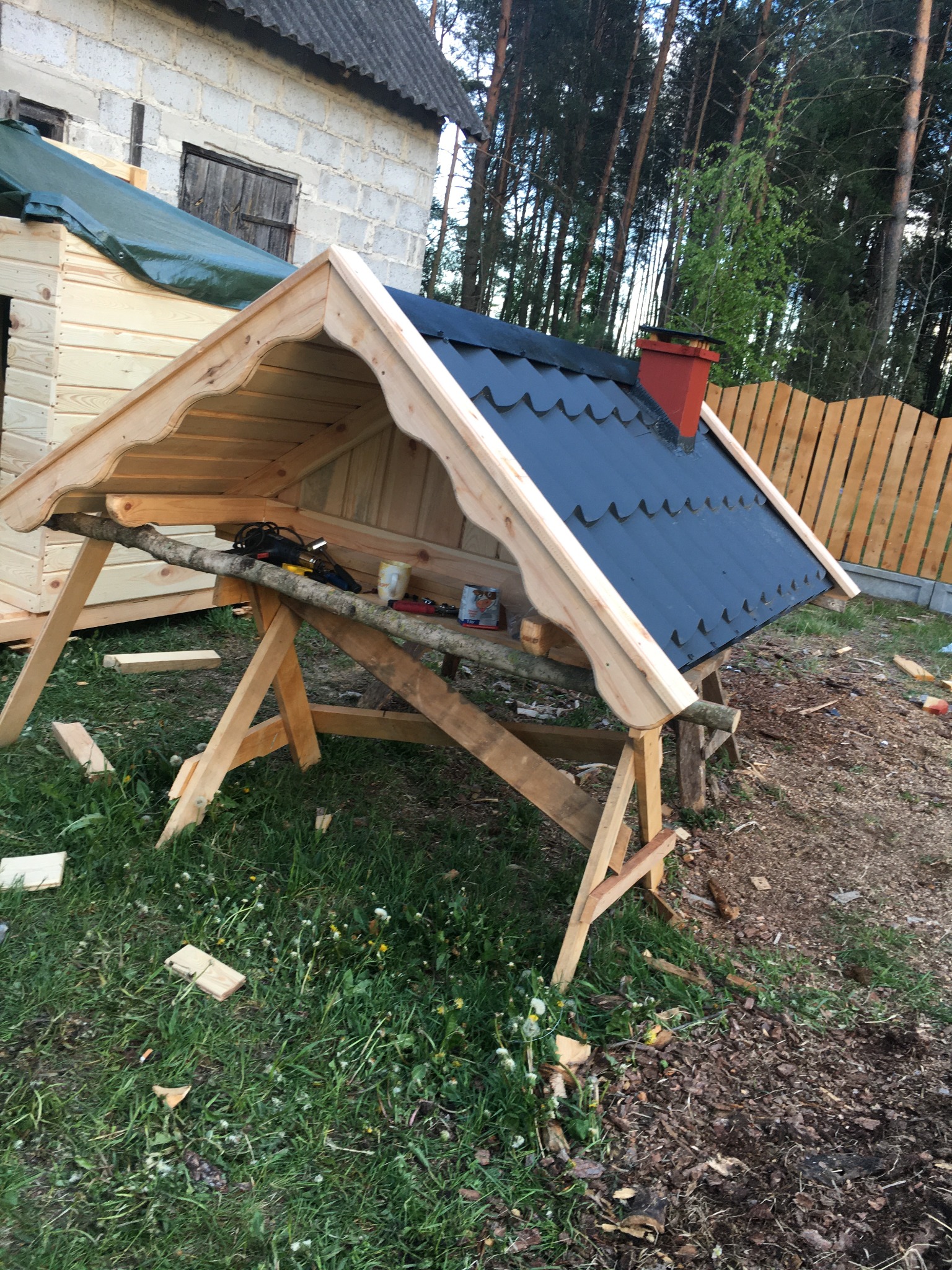 Konstrukcja dachu w trakcie budowy: drewniany szkielet z czarnym pokryciem, czerwony komin, narzędzia na prowizorycznym rusztowaniu, trawa i budynek w tle.