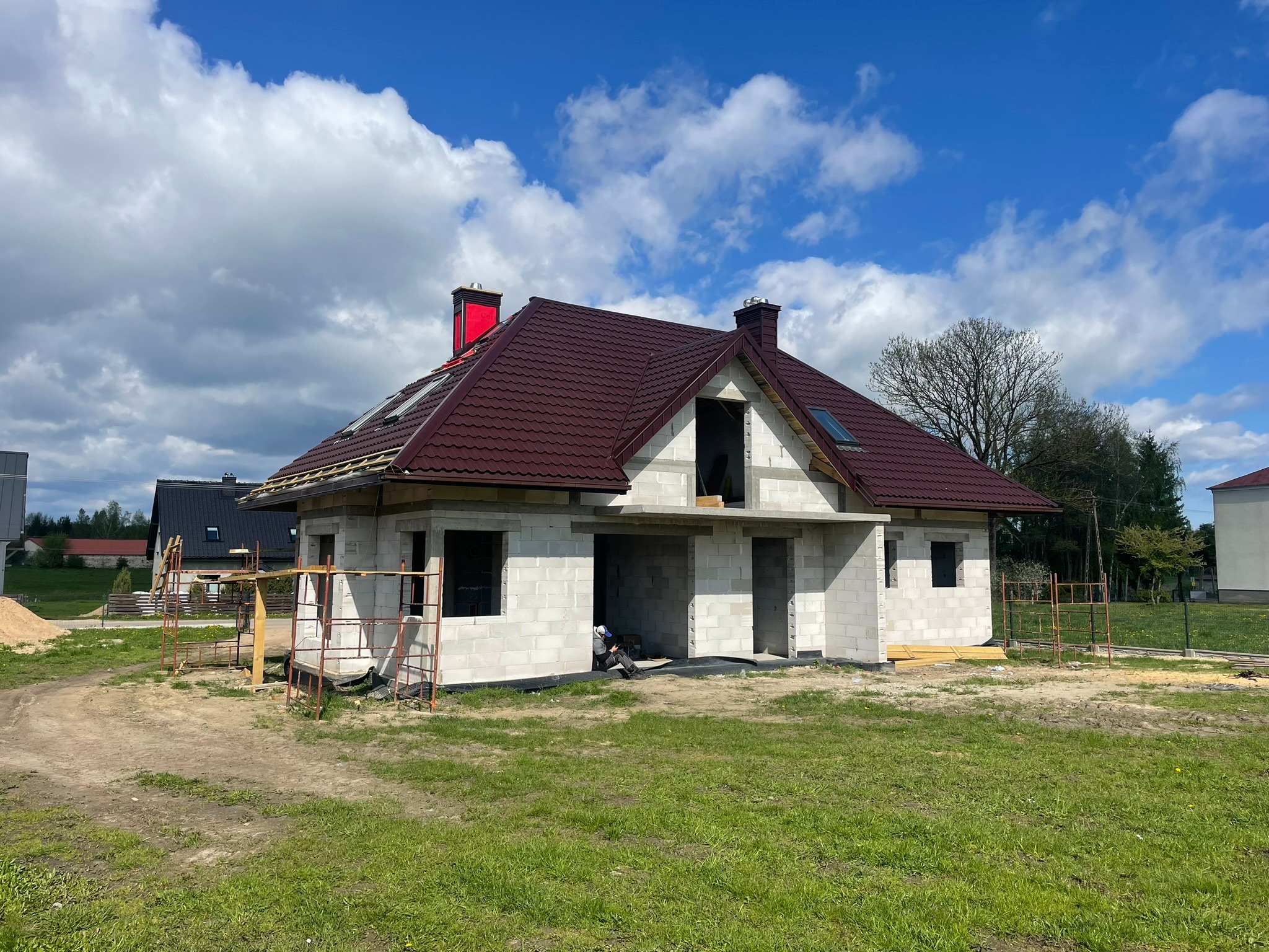 Dom w trakcie budowy z częściowo ukończonym, brązowym dachem z blachy trapezowej i czerwonym kominem. Widoczne rusztowania i elementy konstrukcyjne.
