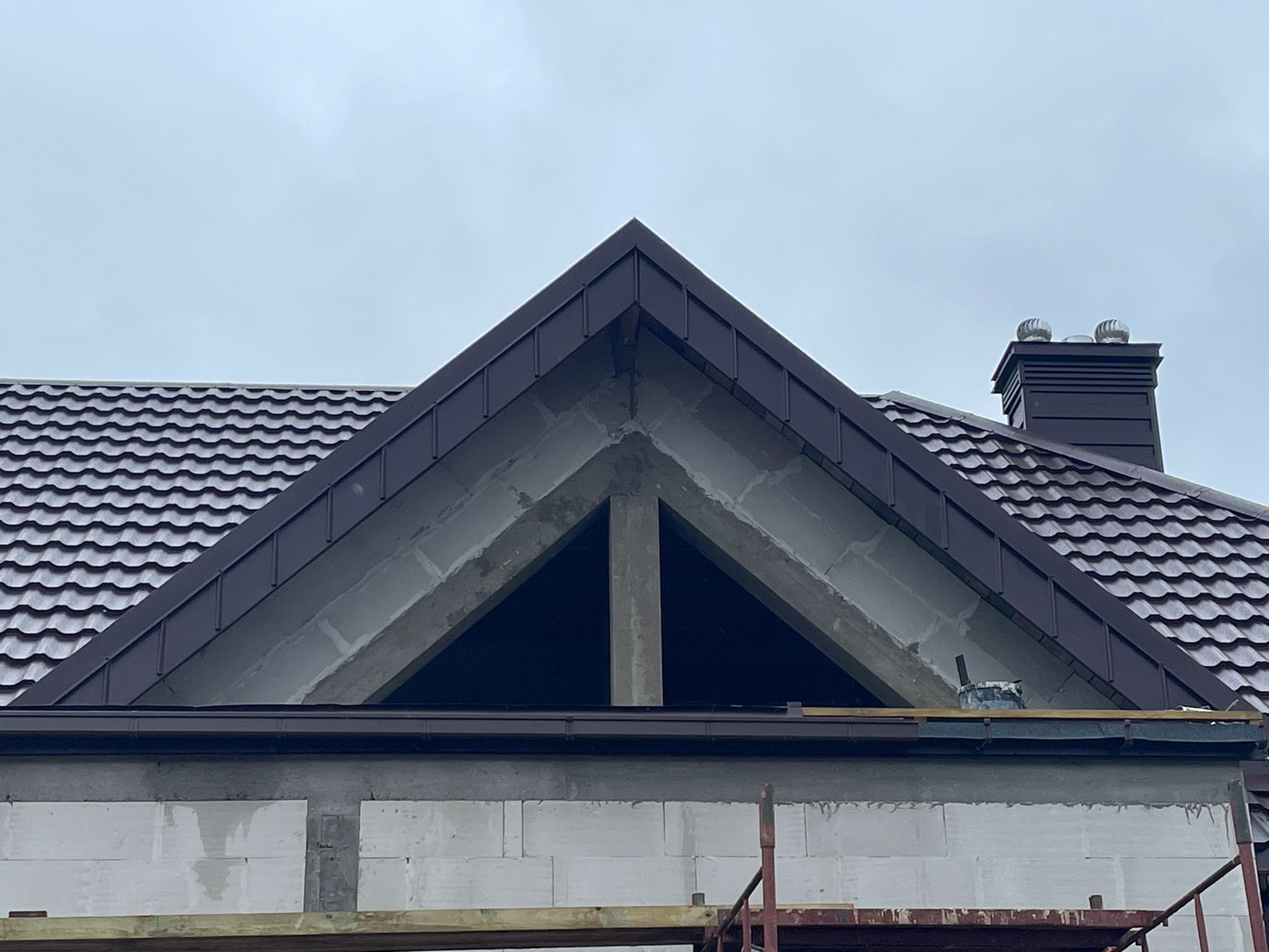 Fragment dachu z brązową blachodachówką i kominem w Jednorożcu, widok z dołu na szczyt ściany z pustaków. Widoczne rusztowanie i elementy konstrukcyjne.