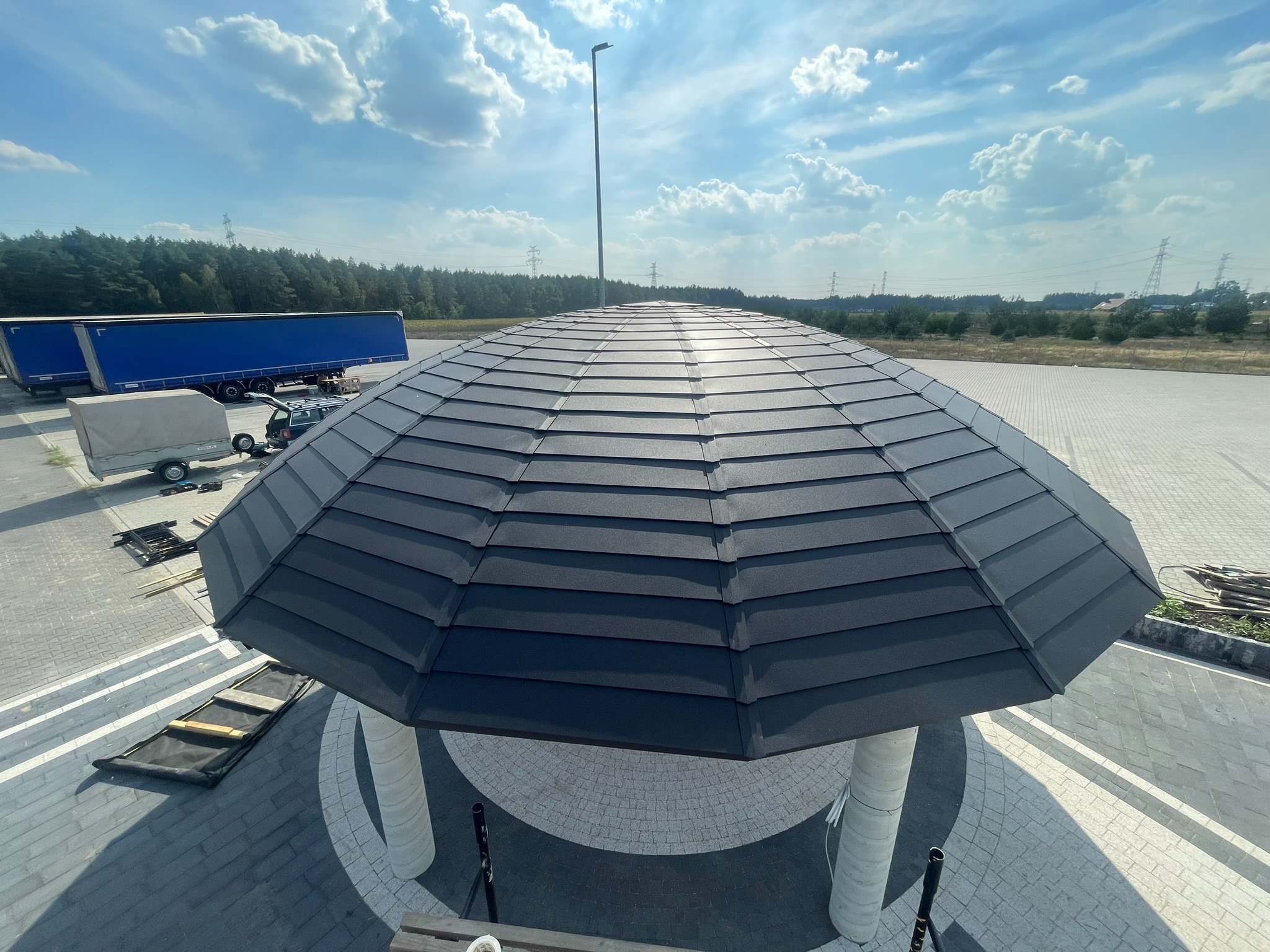 Nowoczesna altana z ciemnoszarą dachówką, widok z góry na tle błękitnego nieba i zieleni. Nietypowy kształt dachu i jasne kolumny altany.