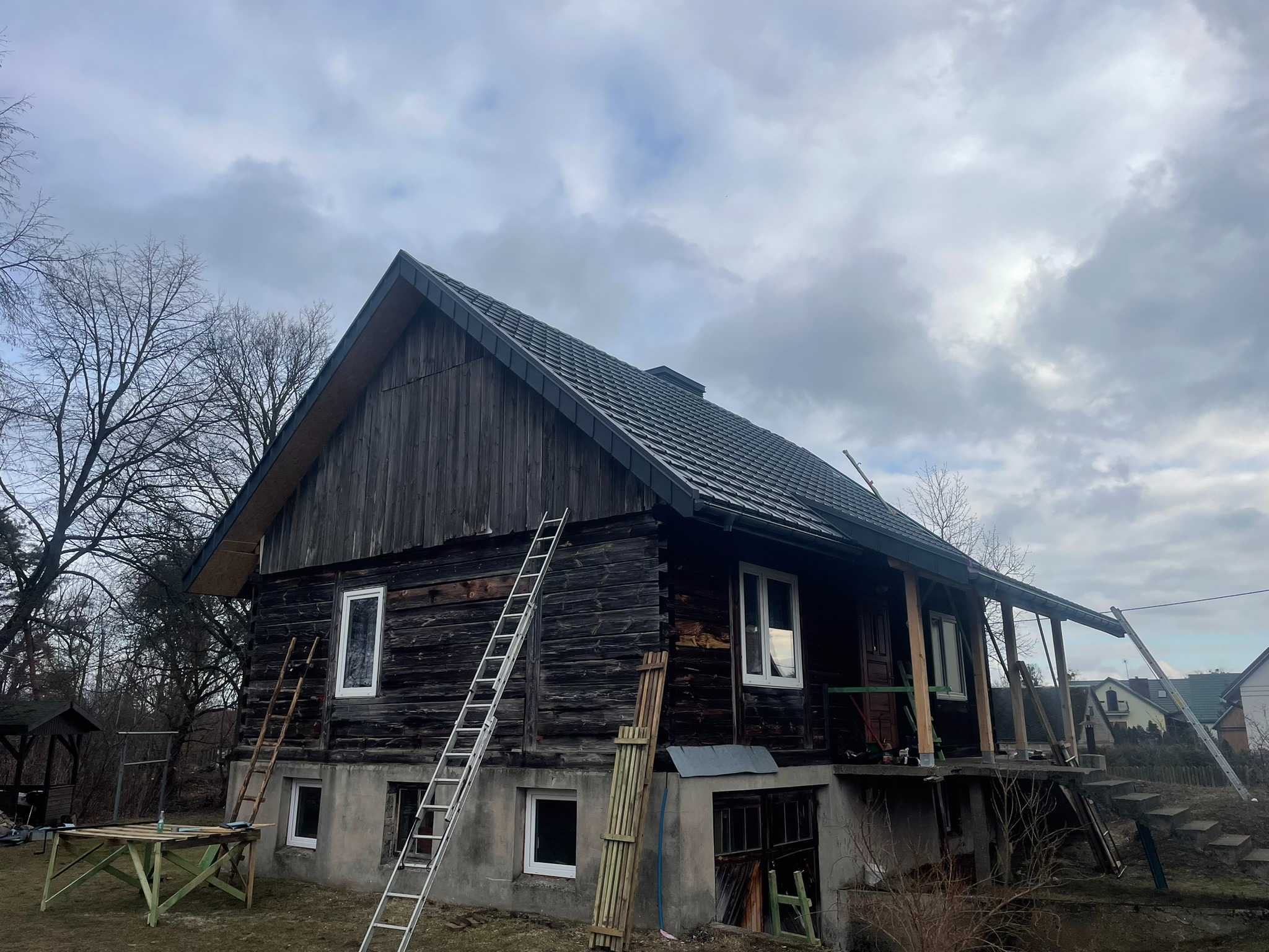 Ciemny, drewniany dom z nowym, grafitowym dachem. Dwie drabiny opierają się o ścianę. Widoczne elementy konstrukcyjne werandy. Pochmurne niebo w tle.