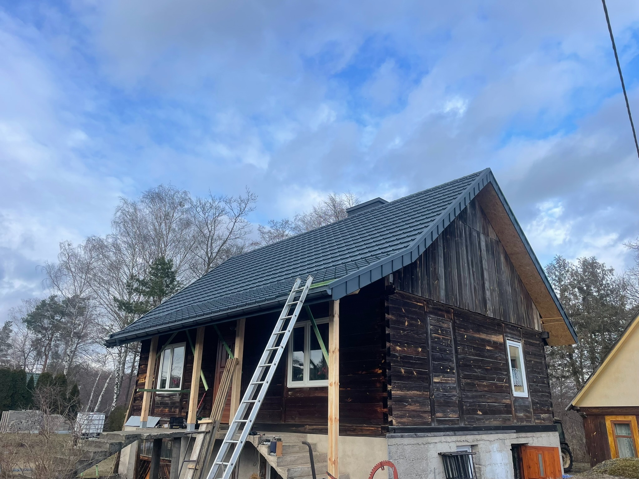 Ciemny, drewniany dom z nowym, grafitowym dachem z blachy na tle pochmurnego nieba. Aluminiowa drabina oparta o ścianę. Widoczne elementy konstrukcyjne werandy.
