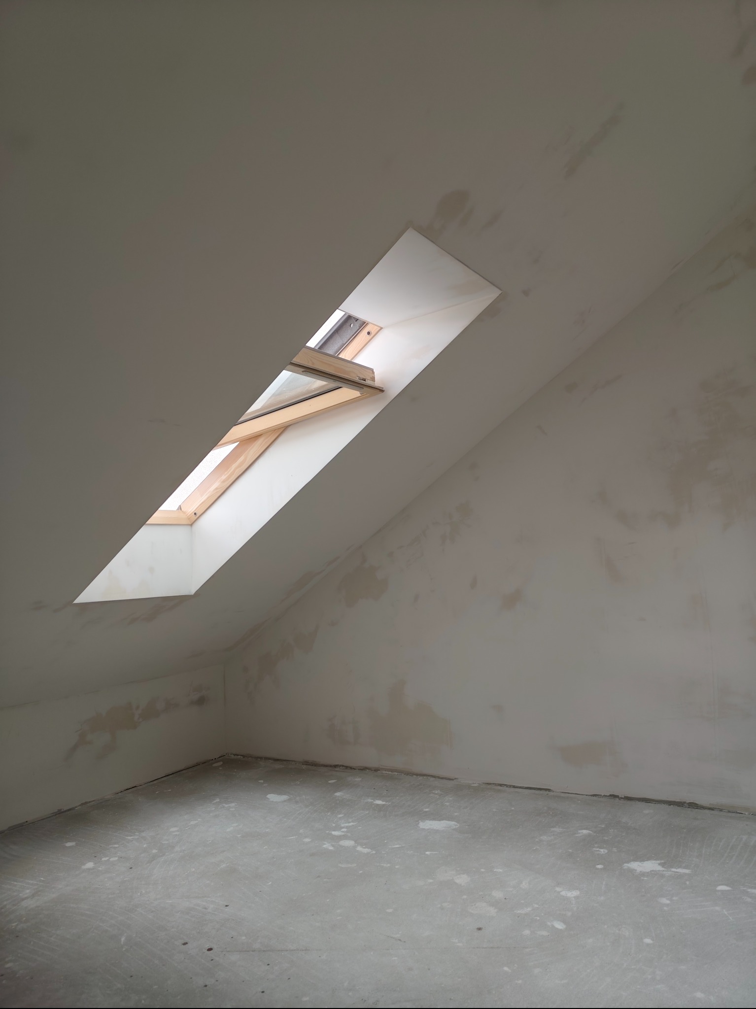 Poddasze ze skośnym oknem dachowym w drewnianej ramie, ściany i podłoga w trakcie szpachlowania, przygotowane do malowania. Widok na surowy stan wykończenia.