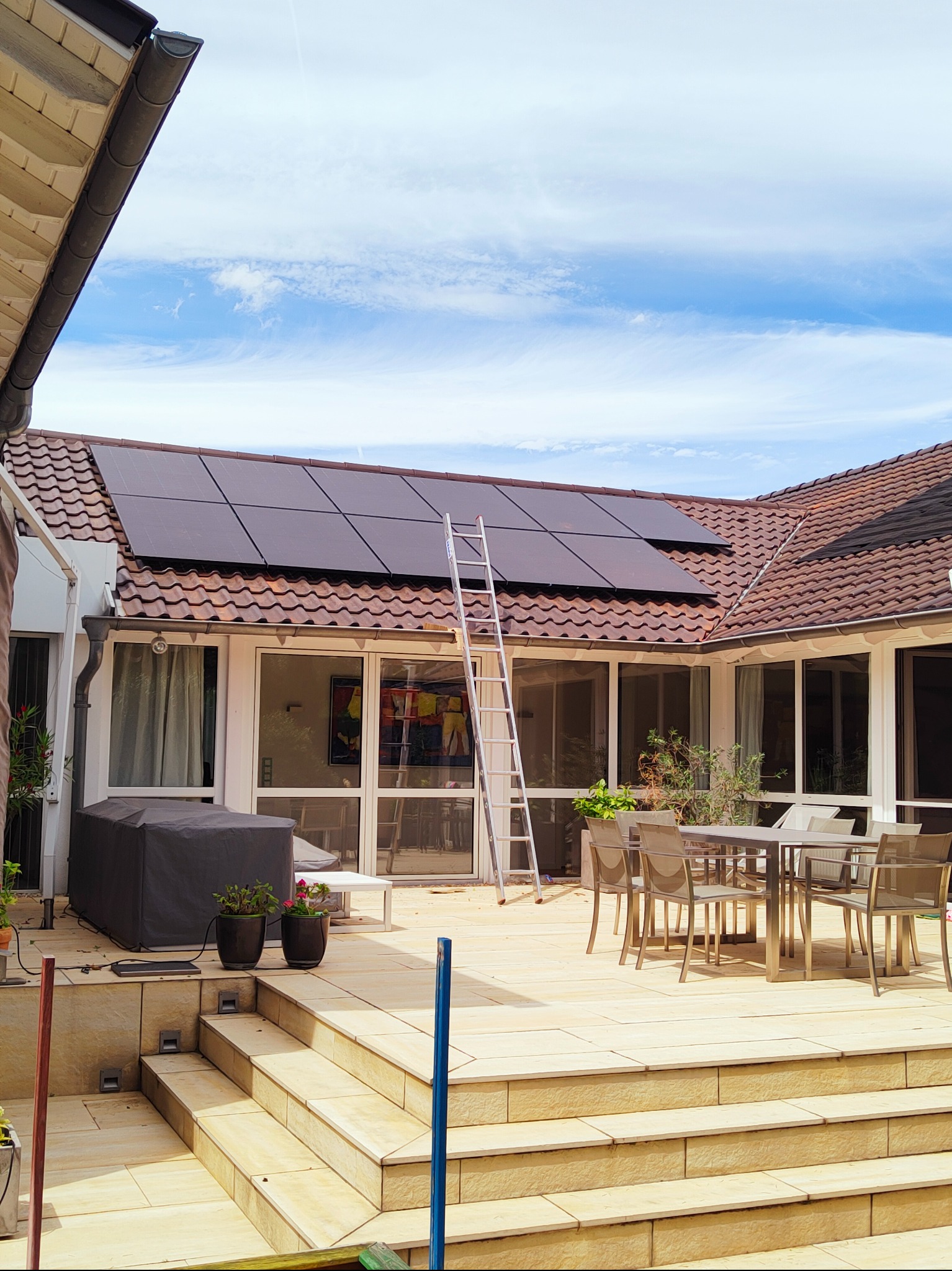 Instalacja paneli słonecznych na dachu domu z tarasem. Aluminiowa drabina oparta o dach. Widok na błękitne niebo z lekkimi chmurami.