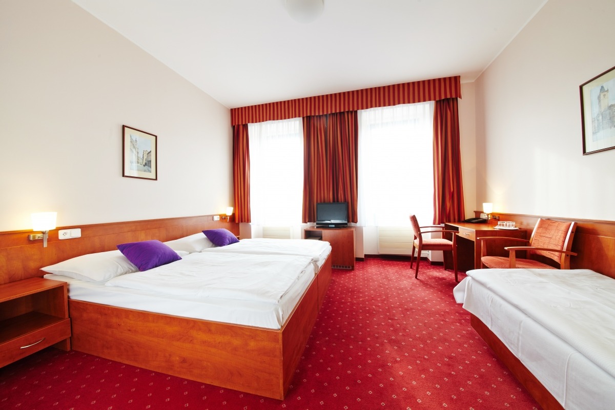 Pokój hotelowy z dwoma łóżkami, biurkiem i czerwonym dywanem. Widoczne okna z czerwonymi zasłonami i obraz na ścianie. Przytulne wnętrze.