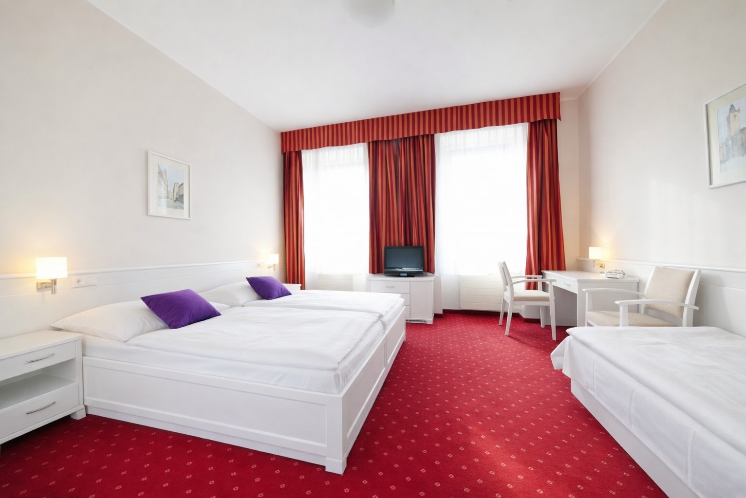 Jasny pokój hotelowy z dwoma łóżkami, biurkiem i czerwonym dywanem. Fioletowe poduszki na łóżku, białe meble i ściany. Okna z czerwonymi zasłonami w tle.