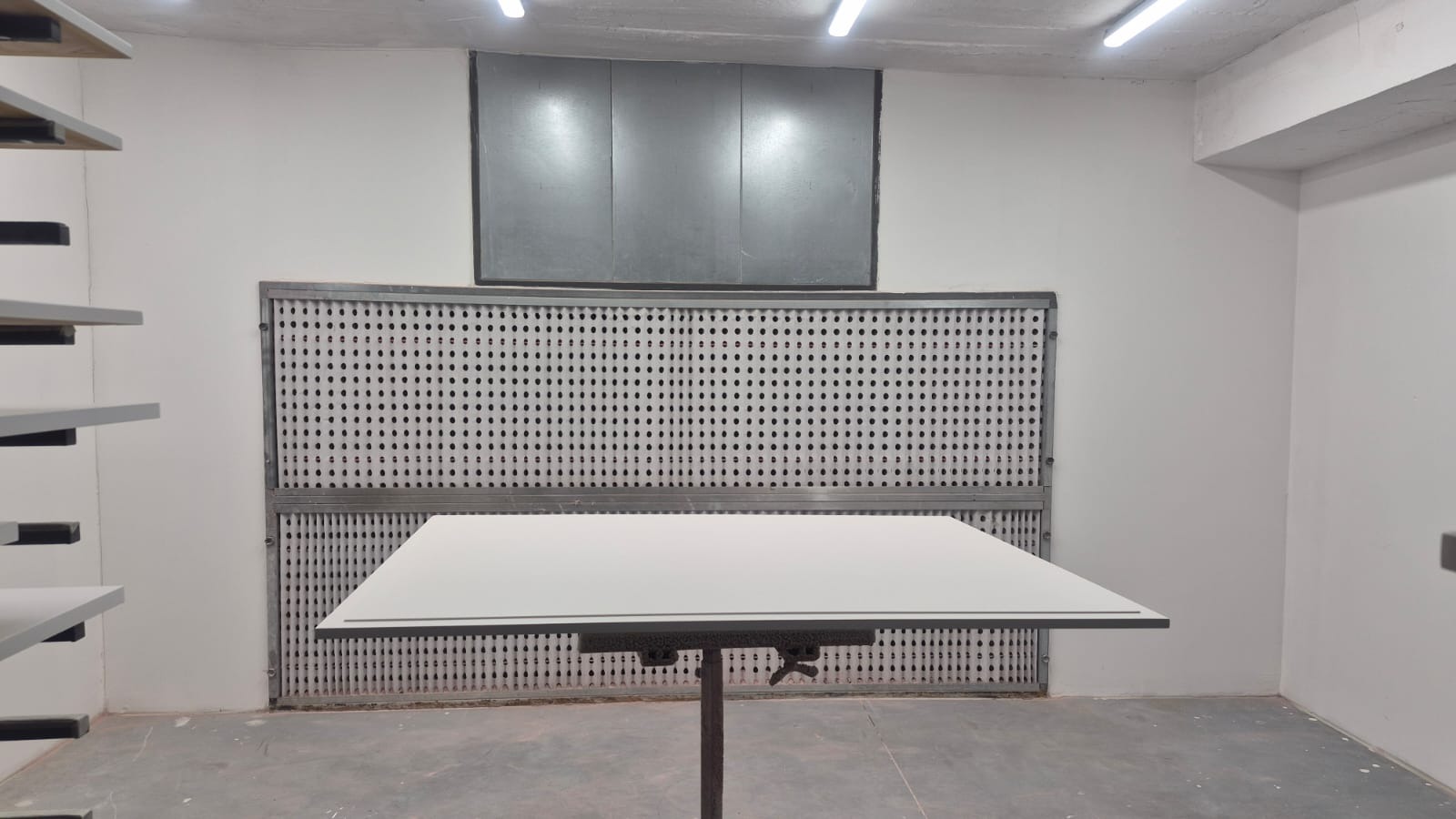 Biały blat stołu na metalowej nodze w lakierni z perforowaną ścianą i oświetleniem sufitowym. Widoczne półki z elementami meblowymi po lewej stronie.