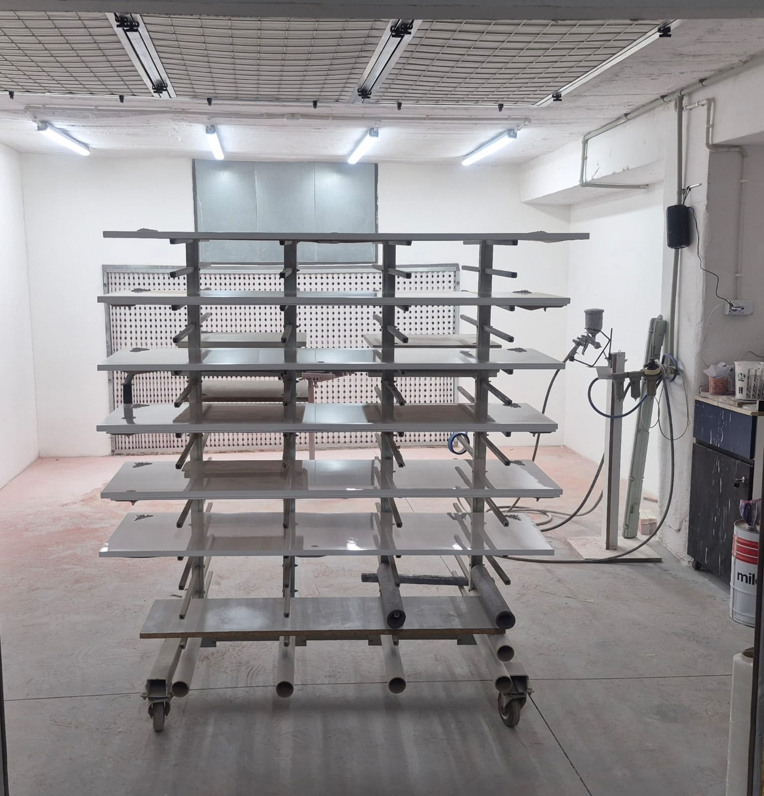 Półki z pomalowanymi na szaro elementami meblowymi w lakierni. Widoczny pistolet lakierniczy i puszki z farbą. Oświetlenie jarzeniowe.