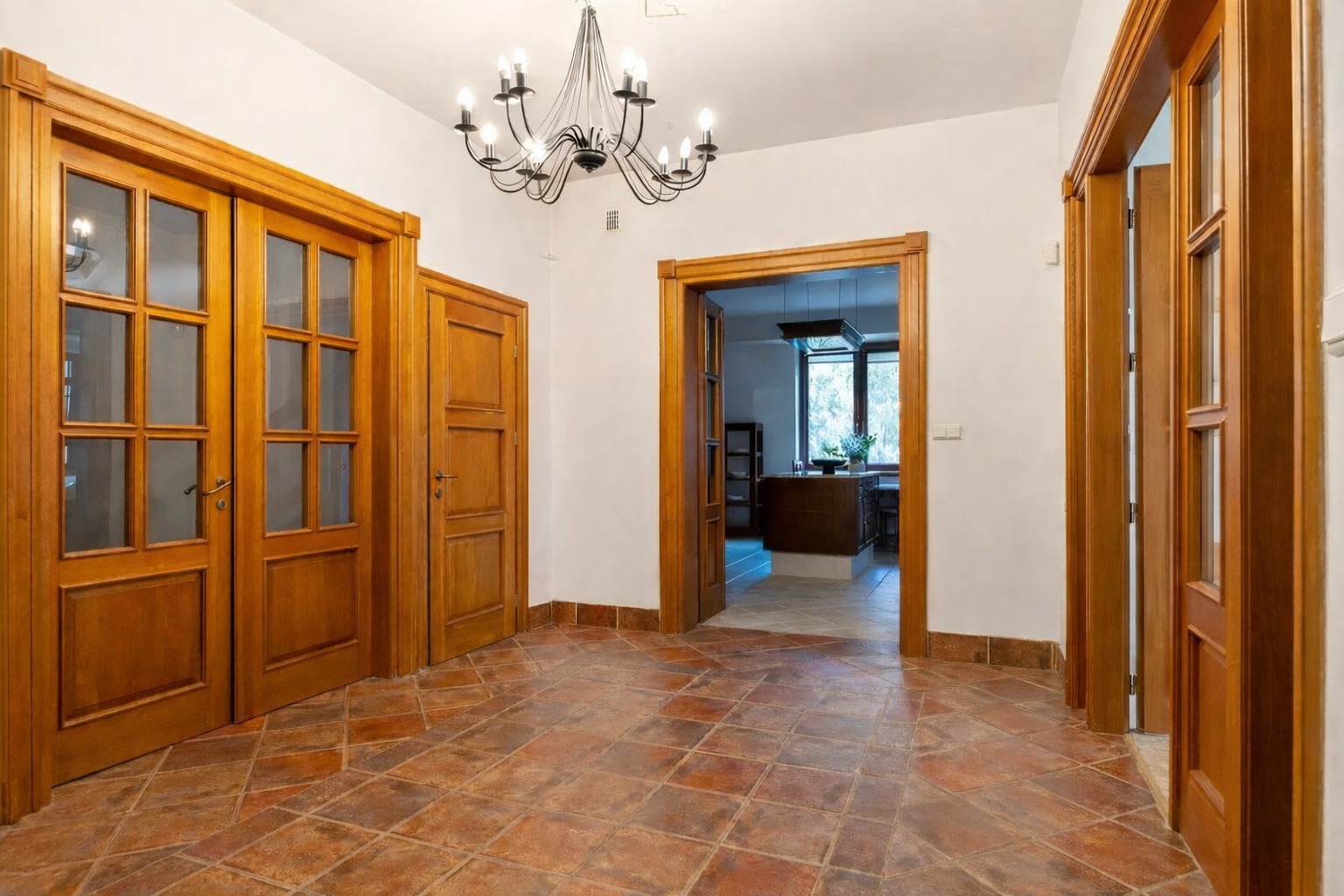 Korytarz z terakotową podłogą i trzema parami drewnianych drzwi. Jedne drzwi są przeszklone, a w tle widać otwartą kuchnię. Nad całością wisi ozdobny, czarny żyrandol.