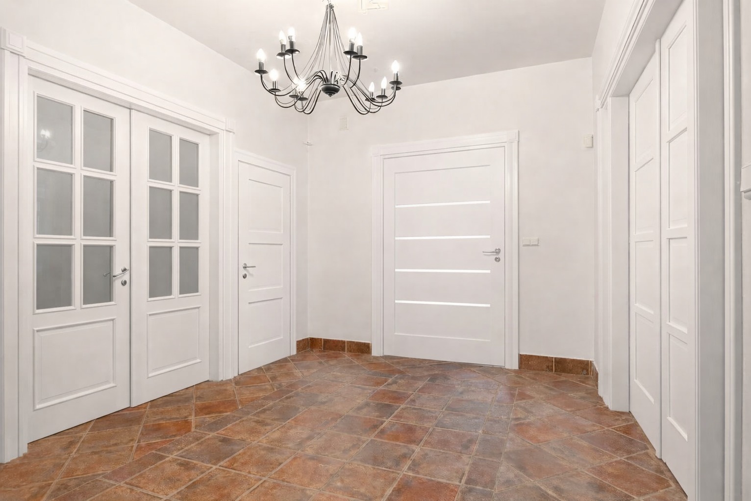 Jasne wnętrze z trzema białymi drzwiami o różnym designie i terakotową podłogą. W tle widoczny żyrandol.
