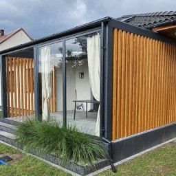 Dtgdrewno - Nowoczesna zabudowa tarasowa z drewna i szkła, z zasłonami bawełnianymi, widok z zewnątrz na tle zielonego trawnika i fragmentu basenu. Detal: pionowe deski.