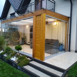 Dtgdrewno - Zadaszenie tarasu z drewna BSH i szkła hartowanego, przesuwne drzwi, widoczne zasłony przeciwsłoneczne, meble ogrodowe wewnątrz, oświetlenie.