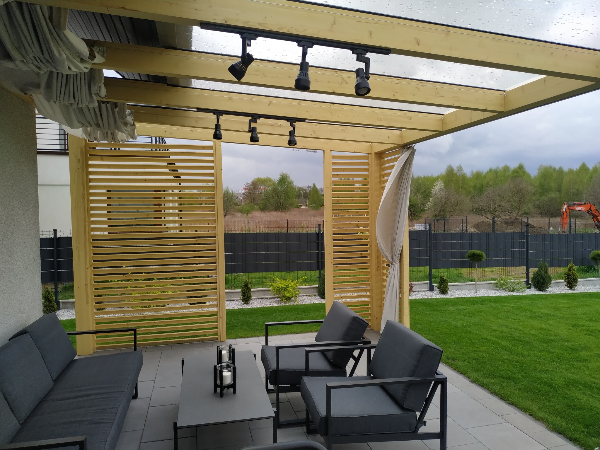 Zadaszony taras z drewna BSH i szkła hartowanego, wyposażony w zasłony przeciwsłoneczne. Widoczne oświetlenie punktowe, meble ogrodowe i zadbany trawnik.