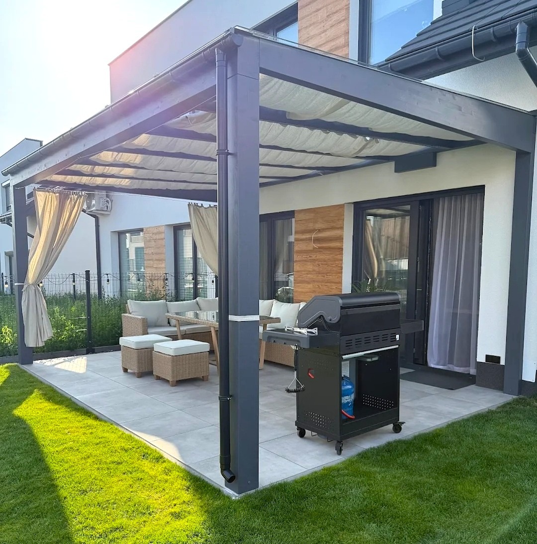 Nowoczesna, szara pergola z zasłonami i szklanym dachem, na tarasie z meblami ogrodowymi i grillem. Widok na zadbany trawnik i dom.