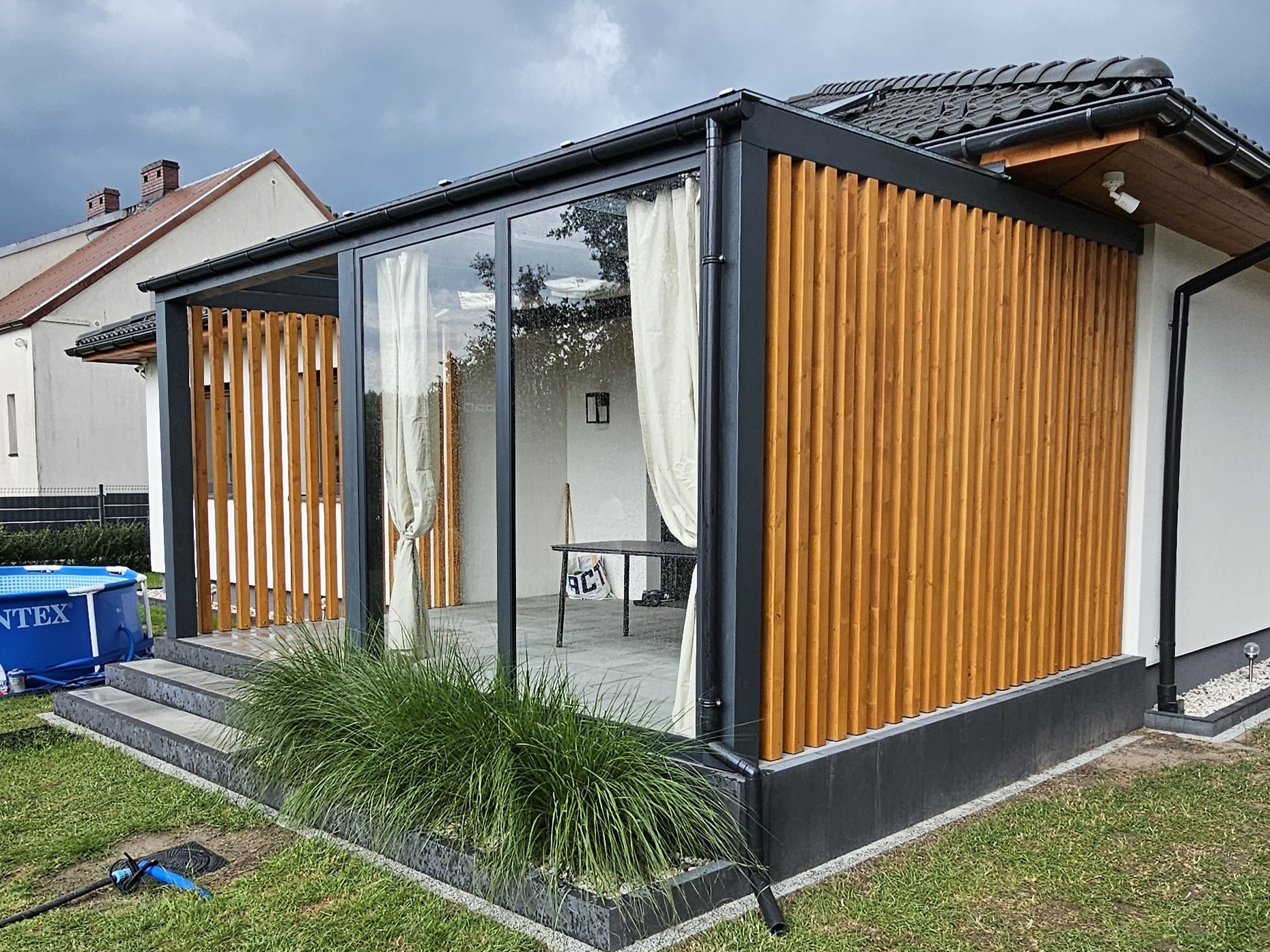 Nowoczesna zabudowa tarasowa z drewna i szkła, z zasłonami bawełnianymi, widok z zewnątrz na tle zielonego trawnika i fragmentu basenu. Detal: pionowe deski.