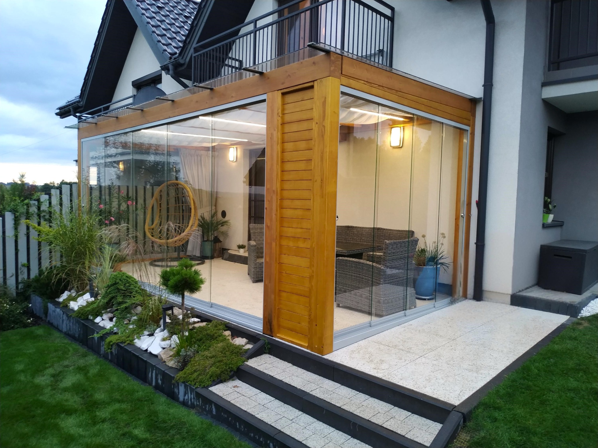 Zadaszenie tarasu z drewna BSH i szkła hartowanego, przesuwne drzwi, widoczne zasłony przeciwsłoneczne, meble ogrodowe wewnątrz, oświetlenie.