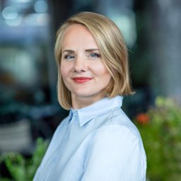 Pośrednictwo Ubezpieczeniowo-Finansowe Małgorzata Ziółkowska - Portret kobiety w błękitnej koszuli, z krótkimi blond włosami i delikatnym makijażem, na rozmytym tle biurowym. Uśmiech, profesjonalny wizerunek.