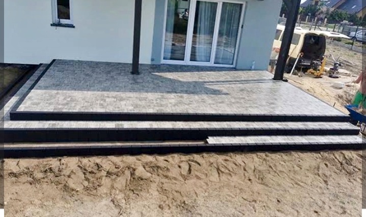 Nowoczesny taras z kostki brukowej w odcieniach szarości, z czarnym obramowaniem, prowadzący do domu z białymi drzwiami balkonowymi. Widoczny fragment ogrodu i sprzęt budowlany.