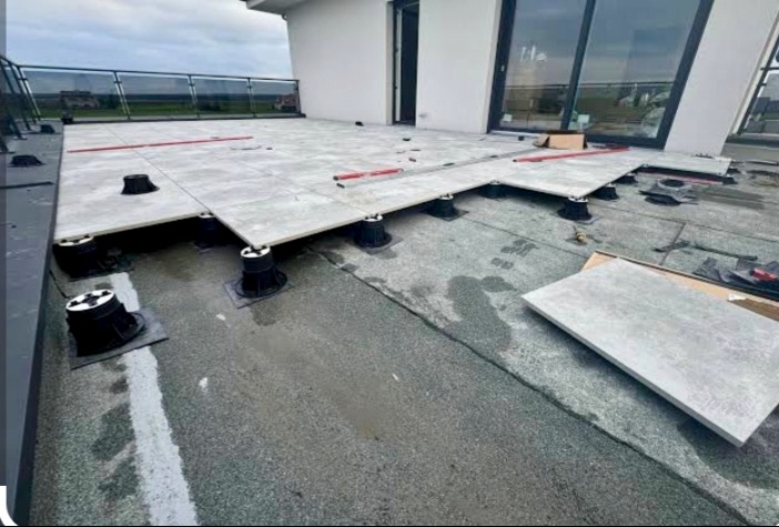 Montaż tarasu na regulowanych wspornikach. Szare płyty ułożone częściowo, widoczne wsporniki i poziomica. Tło: budynek z dużymi oknami i szklana balustrada.