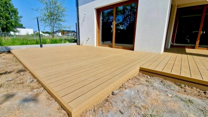 Jasny taras z desek kompozytowych przylegający do domu z dużymi oknami tarasowymi. Widoczne niedokończone prace ziemne wokół tarasu.
