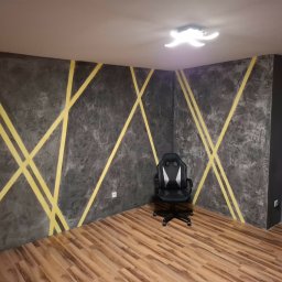 Faint Tech Bud - Pokój po malowaniu ścian z efektem betonu i żółtymi pasami. Na podłodze panele, w rogu krzesło gamingowe. Widoczne gniazdka elektryczne i oświetlenie sufitowe.