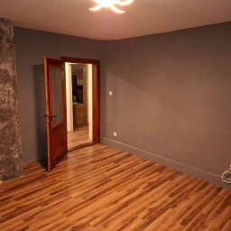 ... - Pokój po remoncie: szare ściany, drewniana podłoga, otwarte drzwi z szybą. Na ścianie dekoracyjny beton z żółtymi pasami. Widoczne gniazdka elektryczne.