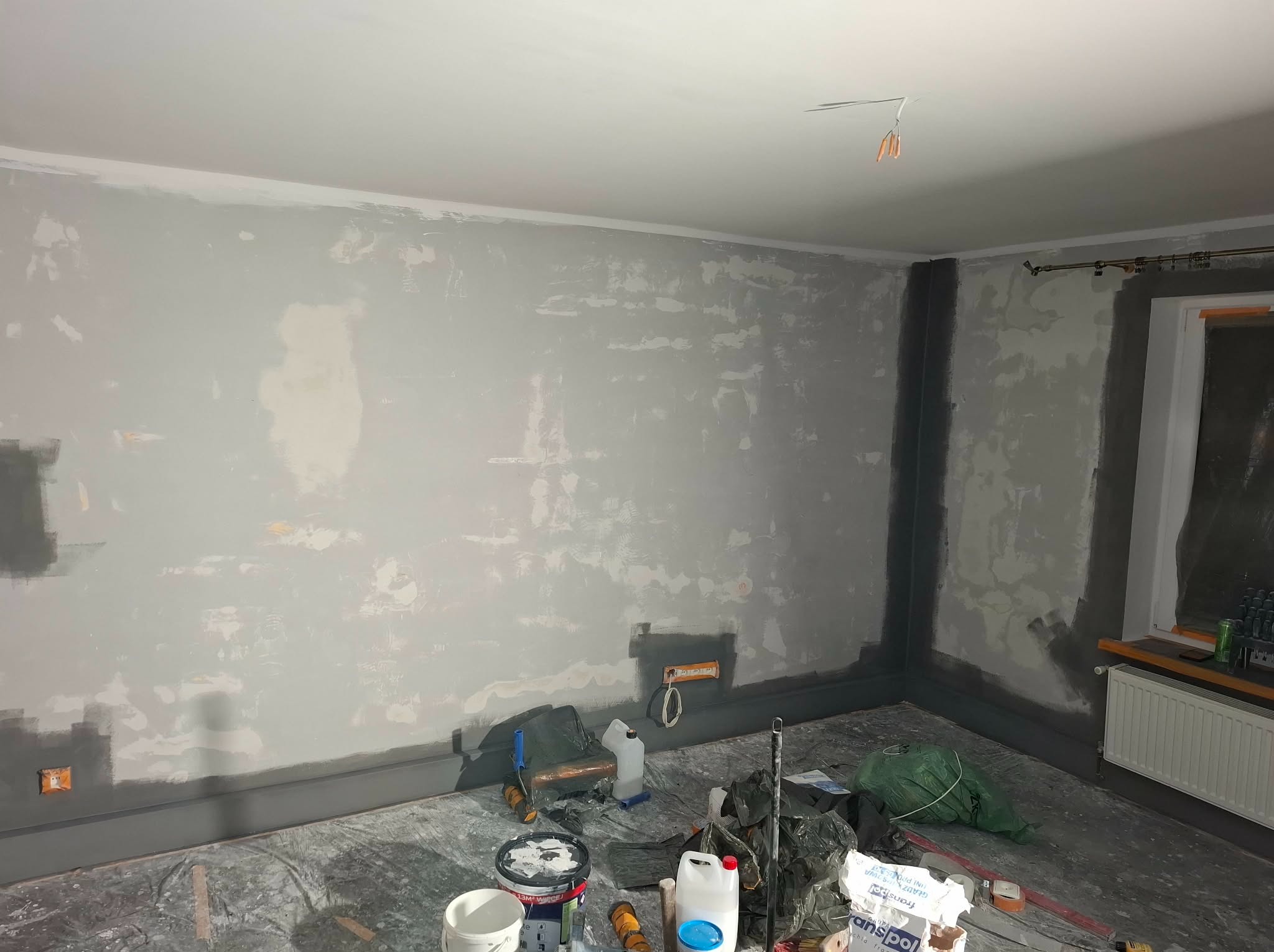 Pokój w trakcie szpachlowania i malowania, ściany częściowo pokryte szarą farbą, widoczne narzędzia i materiały malarskie, folia ochronna na podłodze, grzejnik pod oknem.